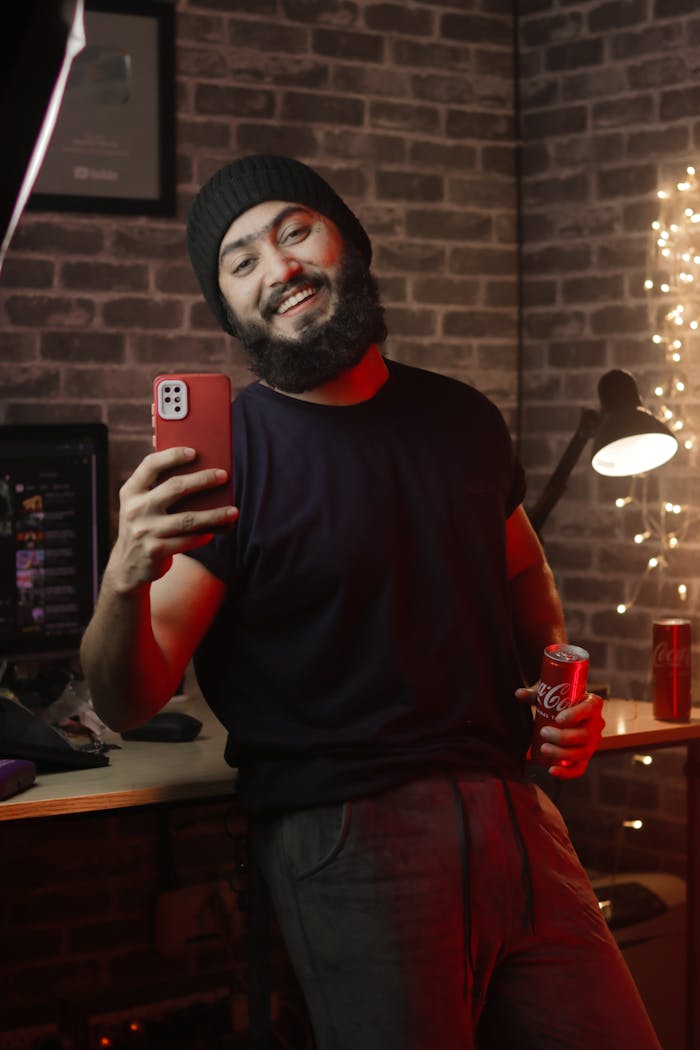 Smiling bearded man taking a selfie with a smartphone, holding a drink, indoors.