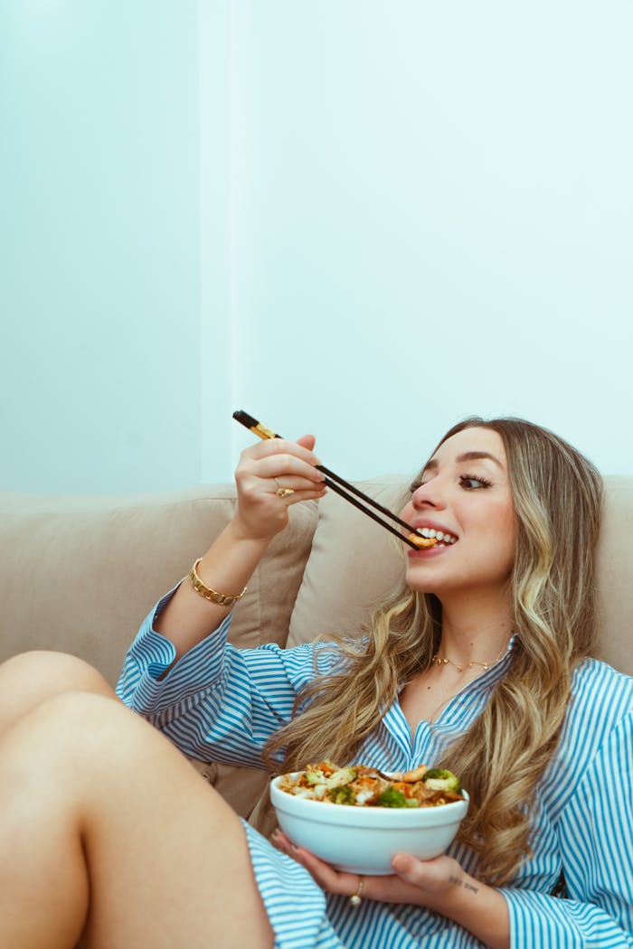 Woman in casual attire eating Asian food with chopsticks on a beige couch.