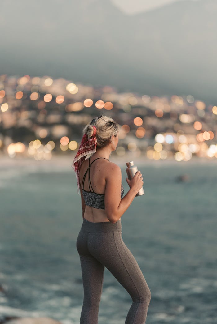 Back view of a woman in activewear holding a tumbler, enjoying the Cape Town beach view during sunset.