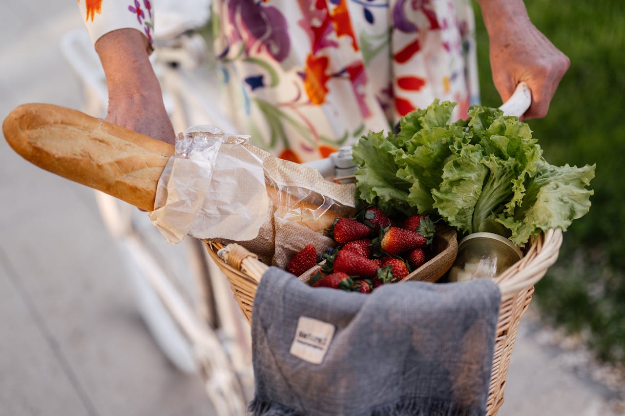A woman in a floral dress holds a basket filled with bread, strawberries, and lettuce on her bicycle.